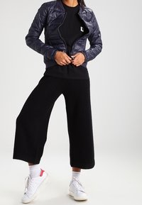 Mörkblå västjacka med dragkedja, svart långärmad topp och vida svarta cropped byxor. Vita sneakers med röda detaljer fulländar outfiten.