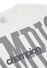 Weißes Baumwoll-T-Shirt mit großen grauen Buchstaben, die "CHAMPION" und "NEW YORK" lesen. Runder Ausschnitt und weicher Stoff.