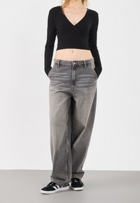 Woman wearing a black long-sleeve cropped top, loose faded gray jeans with hands in pockets, and black sneakers with white stripes.