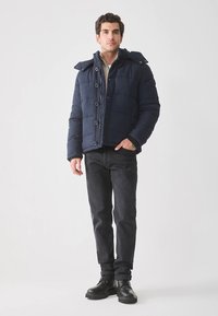 Donkerblauwe gewatteerde jas met capuchon, voorzien van een knoopsluiting en een gestructureerde stof. Gecombineerd met donkere spijkerbroek en zwarte laarzen.