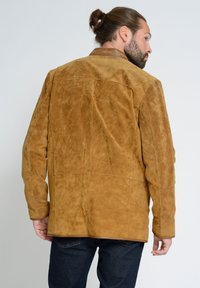 Bruine suède jas met een gestructureerde afwerking, voorzien van een klassieke kraag en schoudernaden, gedragen over donkere jeans.