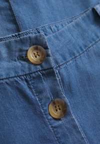 Tissu bleu clair avec une texture lisse, comportant deux boutons ronds transparents à quatre trous, positionnés sur une section froncée.