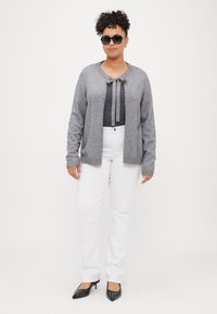 Harmaa neule, jossa on pyöreä pääntie ja etuosassa nauhakoriste, kerroksittain tumman, kimaltelevan topin päällä, yhdistettynä valkoisiin suoriin housuihin ja mustiin niittikorkokenkiin.