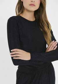 Čierne tričko s dlhým rukávom vyrobené z trblietavého, štruktúrovaného materiálu. Má okrúhly výstrih a priliehavé rukávy, s voľným strihom v oblasti pása.