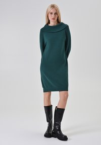 Abito lavorato a maglia verde con colletto sovradimensionato e maniche lunghe, abbinato a stivali in pelle nera al ginocchio con zip laterale.