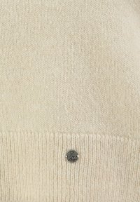 Maglione a maglia color crema con orlo a costine, dotato di un piccolo tag in argento vicino al fondo, che mette in risalto una texture morbida e un design casual.