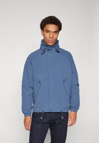 Blue jacket with a high collar, zipper and snap closures, side pockets, and adjustable drawstring at the hem; smooth texture.