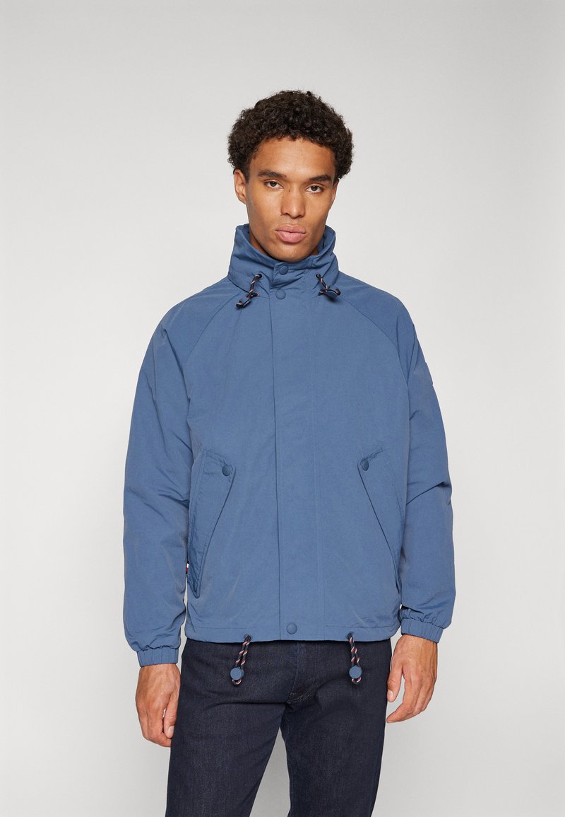 Blue jacket with a high collar, zipper and snap closures, side pockets, and adjustable drawstring at the hem; smooth texture.