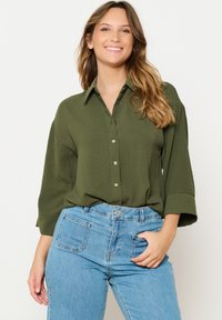 Olive green button-up shirt with a collar, relaxed fit, and three-quarter sleeves, paired with light blue high-waisted jeans.