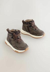 Braune High-Top-Sneaker aus weichem Material mit Schnürung und Reißverschluss. Weiße Zwischensohle und hellbraune Außensohle mit strukturierten Akzenten.