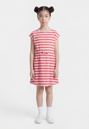 Giovane ragazza in piedi che indossa un vestito a righe rosse e rosa, calzini bianchi e scarpe da ginnastica bianche, con i capelli raccolti in due chignon.