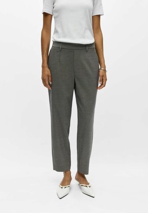 Personne portant un pantalon gris sur mesure, une chemise blanche à manches courtes, des chaussures blanches à bout pointu et un bracelet argenté, debout devant un fond uni.