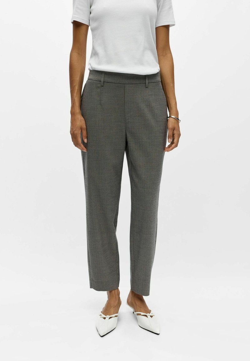 Personne portant un pantalon gris sur mesure, une chemise blanche à manches courtes, des chaussures blanches à bout pointu et un bracelet argenté, debout devant un fond uni.