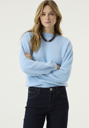 Woman with long blonde hair wearing light blue fuzzy sweater, dark jeans, and dark beaded necklace, standing with arms crossed.