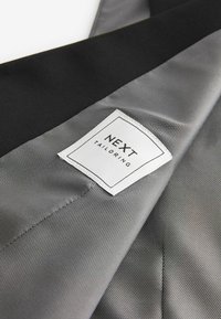 Grau maßgeschneiderter Stoff mit glatter Textur und schwarzem Akzent. Ein weißes Etikett mit der Aufschrift "NEXT TAILORING" ist in den Stoff genäht.