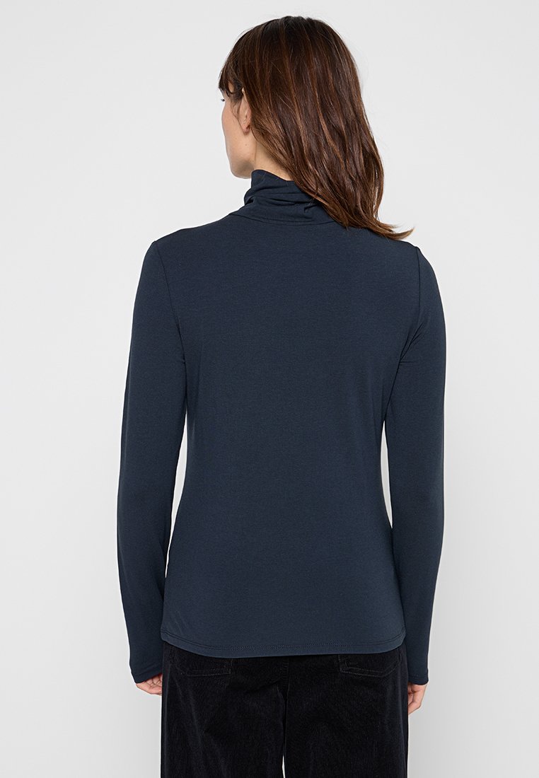 Vrouw met schouderlang bruin haar in een oversized marineblauwe longsleeve coltrui, met haar rug naar de camera gekeerd tegen een effen achtergrond.