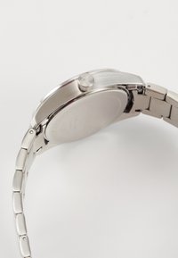 Montre-bracelet en acier inoxydable avec un boîtier poli, une couronne texturée et un bracelet en métal articulé. Le dos est lisse avec des détails gravés.