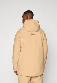 Beige isolert jakke med hette, med svart logo på baksiden, glatt tekstur og avslappet passform. Små detaljer på ermene.