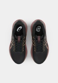 Chaussures de sport noires avec une semelle bordeaux, présentant un matériau texturé, des accents jaunes et un laçage traditionnel. Logo visible sur la languette.