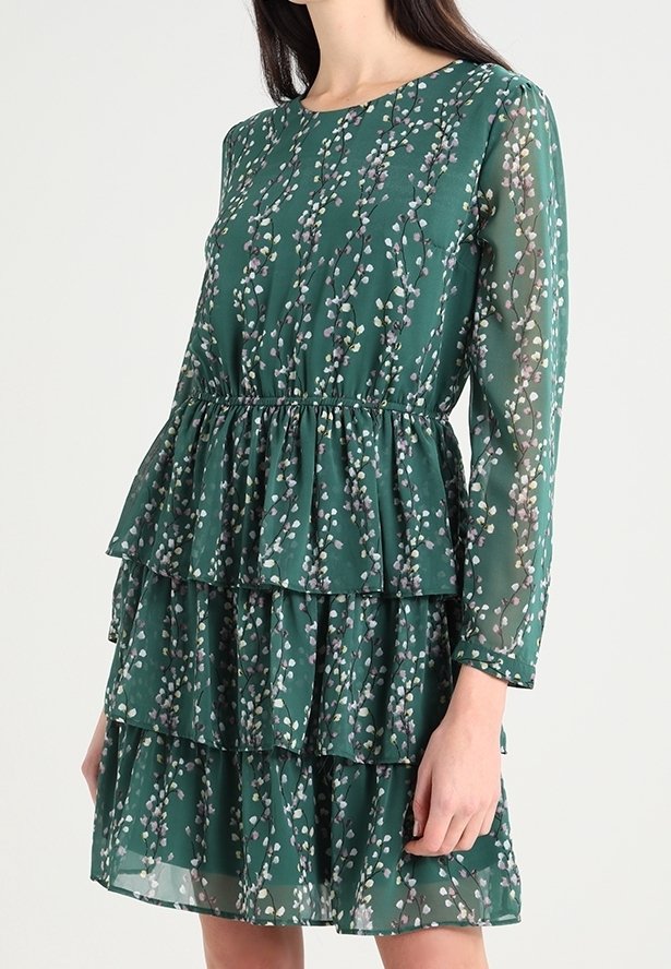 Robe verte à motif floral blanc, manches longues transparentes, jupe à volants à étages et taille froncée, portée par une femme debout.