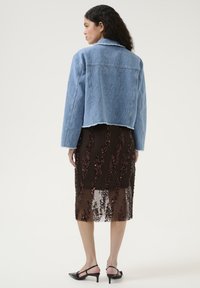 Giacca di jeans cropped azzurro chiaro con orlo sfilacciato abbinata a una gonna trasparente marrone con motivi floreali in paillettes. Scarpe nere con tacco.