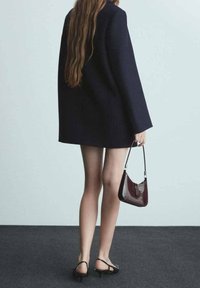 Musta villakangastakki, jossa on leveät hihat, yhdistettynä burgundinväriseen lakatun kankaaseen laukkuun. Malli näyttää jalkojaan ja on pukeutunut mustiin remmissandaaleihin, hiukset valuvat alas.