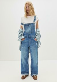 Jonge vrouw staat met haar handen in de zakken, gekleed in een paarse denim overall, een wit T-shirt, een gestreepte blouse om haar armen geknoopt en bruine sneakers.
