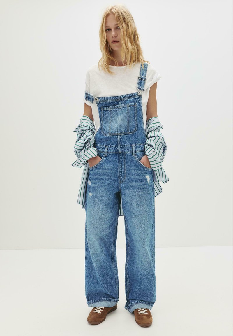 Jonge vrouw staat met haar handen in de zakken, gekleed in een paarse denim overall, een wit T-shirt, een gestreepte blouse om haar armen geknoopt en bruine sneakers.