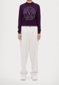 Pull en maille violet avec un logo au centre, associé à un pantalon blanc à fines rayures, présentant une coupe décontractée et des ourlets retroussés. Des baskets blanches complètent le look.
