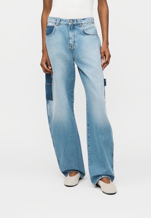 Person, der locker sitzende, hellblaue Jeans aus Denim trägt mit dunkelblauen Aufsatztaschen an den Seiten, kombiniert mit weißen Slip-on Schuhen und einem weißen Oberteil.