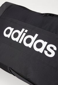 Czarna torba z tkaniny z dużym białym logo "adidas", zamykana na zamek, z elementami teksturowanymi.