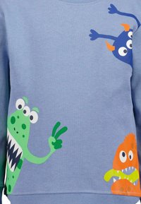 Felpa azzurra chiara con illustrazioni di mostri colorati in verde, blu e arancione, con espressioni facciali e texture distintive.