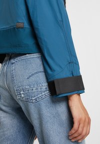 Personne portant une veste bleue avec une sangle noire au niveau du poignet et un jean en denim bleu clair, main détendue le long du corps, étiquette G-Star Originals visible sur la veste.