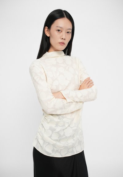 Blusa a maniche lunghe di colore crema con un motivo floreale testurizzato, collo alto, tessuto morbido e una leggera lucentezza; abbinata a una gonna scura.