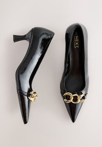 Crne salonke od lakirane kože s oštrim vrhovima. Zlatni lanac na vrhu. Glatka tekstura s sjajnim završetkom, s potpeticom srednje visine.