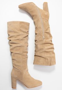 Botas de ante beige hasta la rodilla con diseño fruncido, puntera cuadrada y tacón grueso. Presentan una cremallera lateral para facilitar su uso.
