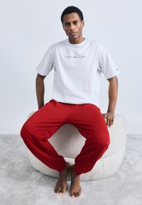 Γκρι βαμβακερό μπλουζάκι με το λογότυπο "TOMMY HILFIGER" σε μαύρο, συνδυασμένο με κόκκινο παντελόνι φόρμας. Το μοντέλο κάθεται σε μια υφασμάτινη μπεζ σκαμπό, ξυπόλητο.