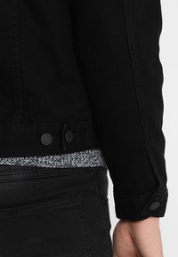 Svart denimjacka med krage, med två svarta knappar i midjan. Bärs över en stickad topp med svart och vitt mönster.