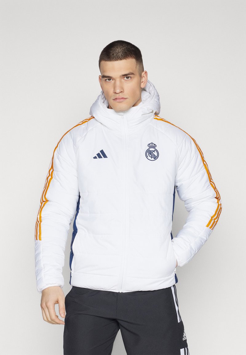 Giacca imbottita bianca con accenti blu navy, strisce arancioni sulle maniche e un alto colletto. Presenta il logo del Real Madrid e il marchio Adidas.