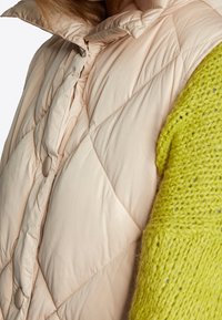 Puffige beige Weste mit Rautenmuster und hohem Kragen, kombiniert mit einem lebhaft grünen Strickpullover mit lockerer Textur.