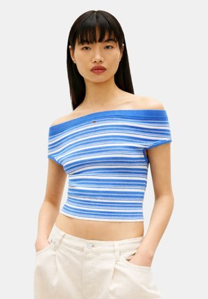 Giovane donna che indossa un top corto a righe orizzontali bianche e blu, scollato sulle spalle, e pantaloni beige chiaro, con le mani in tasca davanti a uno sfondo neutro.