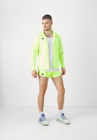 Neon grøn træningsjakke over hvid tanktop, parret med matchende shorts og grå sneakers. Har logo detaljer og letvægtsstof.