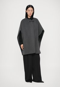 Sivi vuneni plašt s otvorenim prednjim dijelom, s crnim zatvaračem na kopčanje. Nošen preko teksturirane crne bluze i širokih crnih hlača.