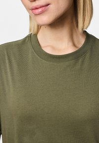 Lähivaade inimesest, kes kannab oliivrohelist kaelusega t-särki, näidates kaela, alumist näoosa ja lühikesi blondi juukseid.