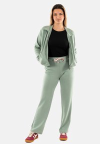 Lichtgroene zip-up jas met ribbels aan de manchetten, gedragen met bijpassende joggers met een trekkoord. Zwarte shirt eronder en multicolor sneakers.