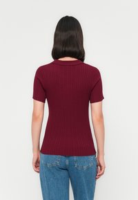Persona con capelli scuri di lunghezza media che indossa una maglietta a coste a maniche corte di color bordeaux e jeans in denim blu, vista da dietro.