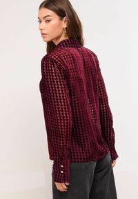 Bordo samta krekls ar houndstooth rakstu, garām piedurknēm ar pogām pie aproces un augstu apkakli, attēlots no aizmugures.