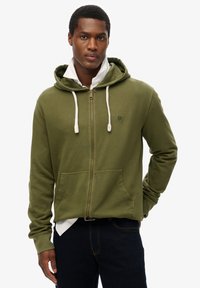 Sudadera con cremallera de color verde oliva, con capucha ajustable, bolsillos frontales, puños acanalados y un pequeño logo bordado en el pecho.