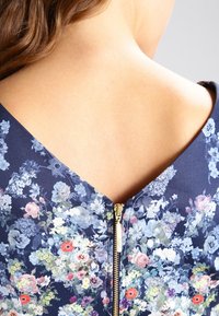 Blumenkleid mit V-Ausschnitt und goldener Reißverschlussöffnung auf der Rückseite. Der Stoff hat einen dunkelblauen Hintergrund mit bunten Blumenmustern.