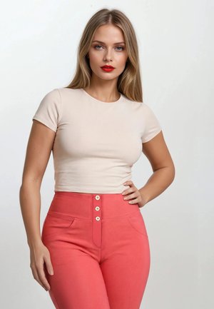 Pitkähiuksinen vaaleahiuksinen nainen pukeutuneena istuvaan beigeen lyhythihaiseen toppiin ja korkeavyötäröisiin korallinvärisiin housuihin, joissa on kolme nappia, poseeraa käsi lanteilla.